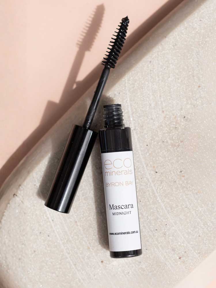 An open tube of Eco Minerals Natural Mascara 8g in the shade Midnight lies on a light textured surface, with the smudge-proof wand angled across the tube.