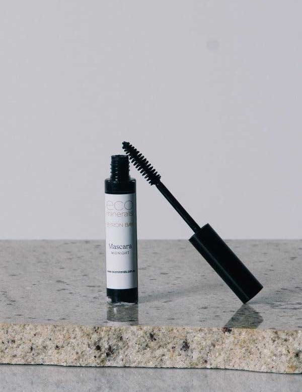 An Eco Minerals Natural Mascara 8g tube with the wand partially removed stands on a speckled stone surface against a plain light gray background.