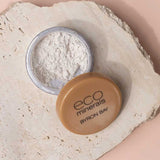 Open container of Eco Minerals White Light Illuminate 5g, a natural mineral highlighter with tan lid, resting on textured beige stone surface—ideal for anyone seeking an eco-friendly makeup option.