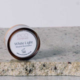 A small round container labeled Eco Minerals White Light Illuminate 5g, a natural highlighter, rests on a textured stone surface with white powder nearby.