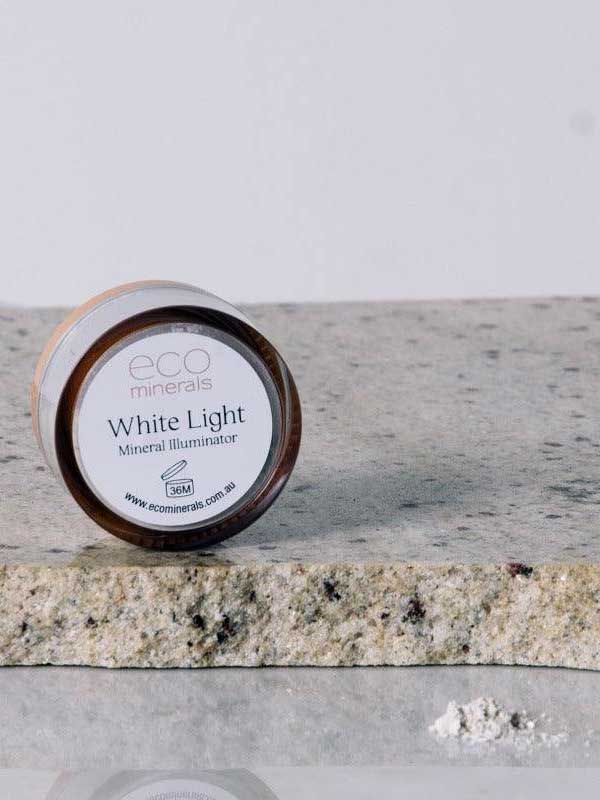 A small round container labeled Eco Minerals White Light Illuminate 5g, a natural highlighter, rests on a textured stone surface with white powder nearby.