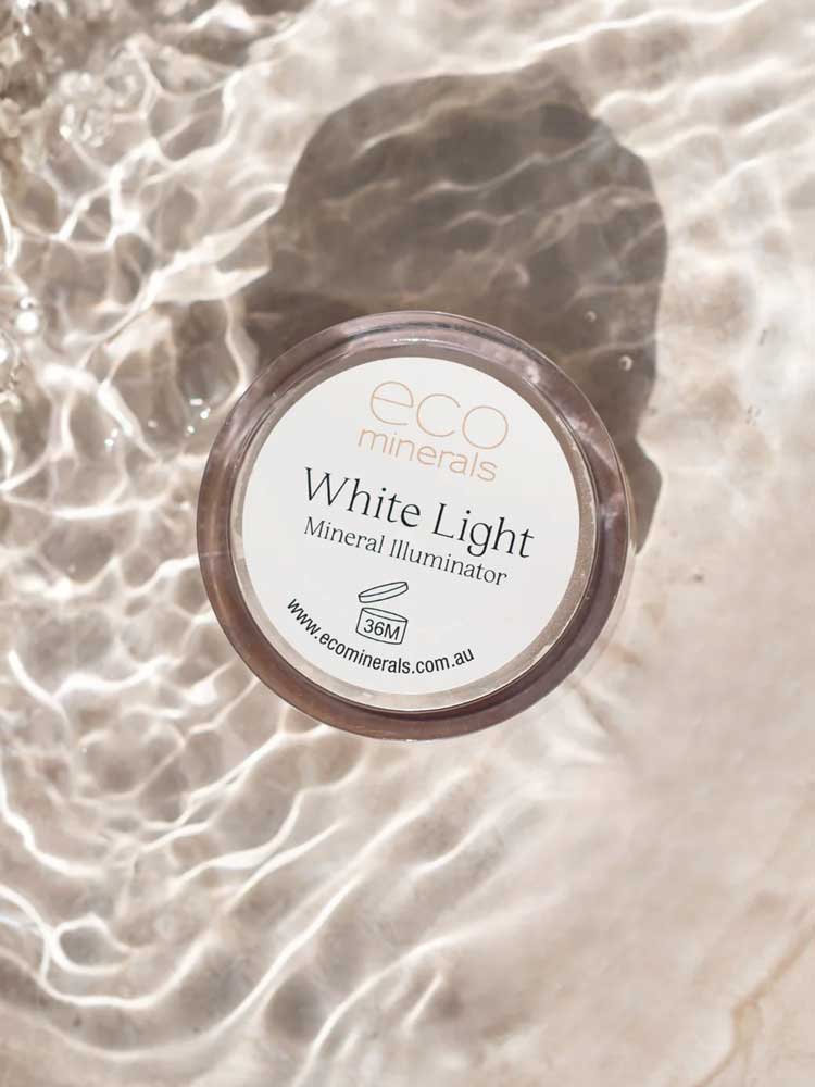 A 5g Eco Minerals White Light Illuminate rests on a light surface with rippling water, casting reflections. The label and website are clearly visible on the lid.