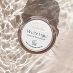 A 5g Eco Minerals White Light Illuminate rests on a light surface with rippling water, casting reflections. The label and website are clearly visible on the lid.