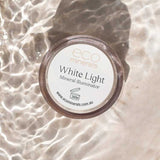 A 5g Eco Minerals White Light Illuminate rests on a light surface with rippling water, casting reflections. The label and website are clearly visible on the lid.