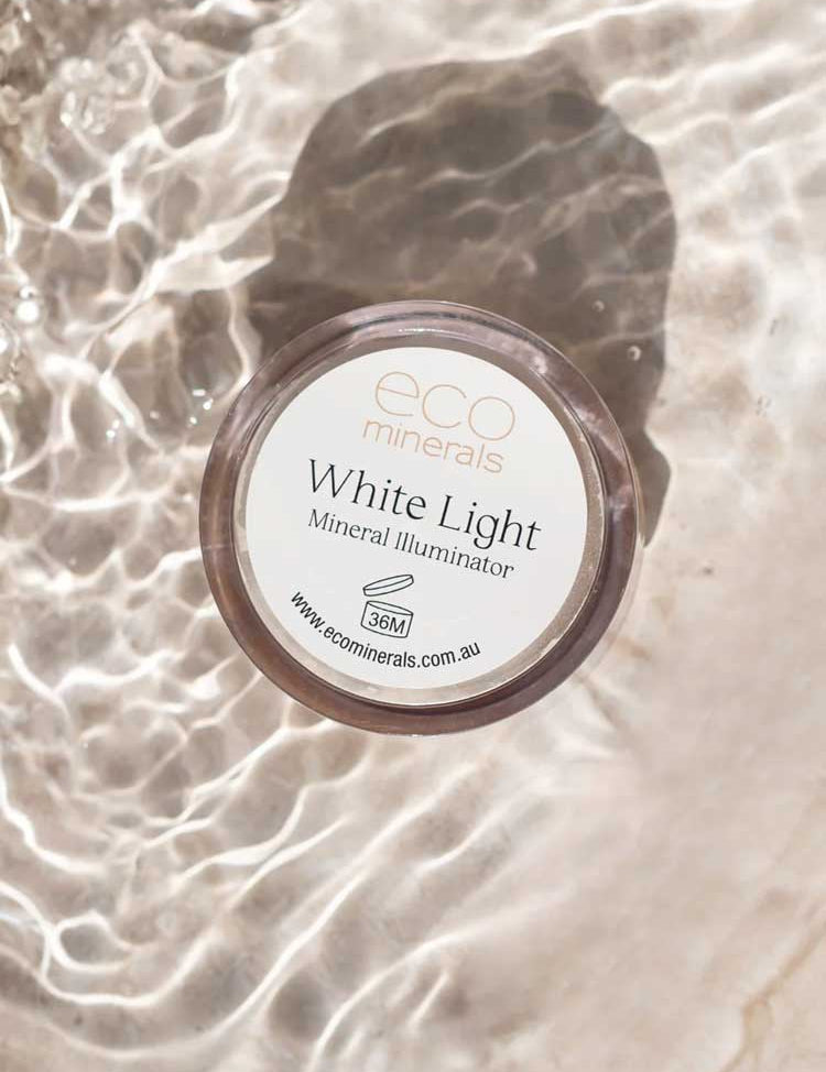 A 5g Eco Minerals White Light Illuminate rests on a light surface with rippling water, casting reflections. The label and website are clearly visible on the lid.