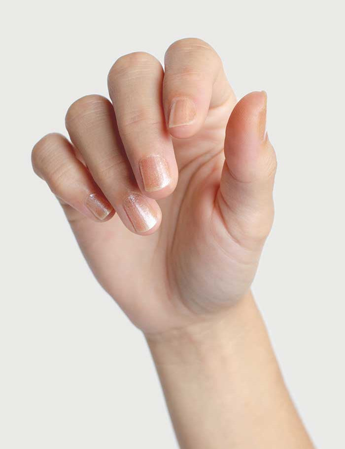 A close-up of a left hand with fingers slightly bent, nails painted in a nude shade using Sienna Byron Bay Nail Polish - Embrace. The background is plain and light gray.