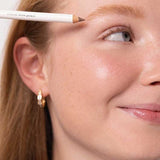 A close-up of a smiling fair-skinned woman with light blonde hair holds Ere Perez Almond Brow Pencil 1.1g near her brow. She wears a gold hoop earring. Text reads: polished | light to medium blonde | vegan brow pencil.