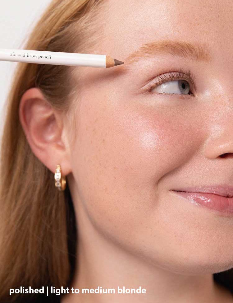 A close-up of a smiling fair-skinned woman with light blonde hair holds Ere Perez Almond Brow Pencil 1.1g near her brow. She wears a gold hoop earring. Text reads: polished | light to medium blonde | vegan brow pencil.