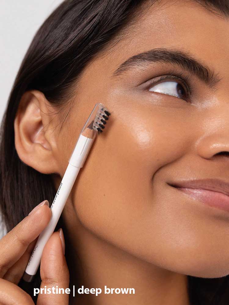A woman with smooth, glowing skin brushes her eyebrow using Ere Perez Almond Brow Pencil 1.1g. The image highlights her face and brows, with the text “pristine | deep brown” displayed at the bottom.