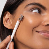 A woman with smooth, glowing skin brushes her eyebrow using Ere Perez Almond Brow Pencil 1.1g. The image highlights her face and brows, with the text “pristine | deep brown” displayed at the bottom.