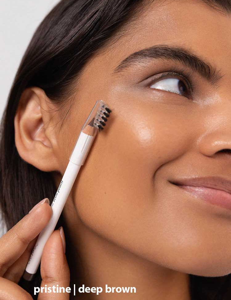 A woman with smooth, glowing skin brushes her eyebrow using Ere Perez Almond Brow Pencil 1.1g. The image highlights her face and brows, with the text “pristine | deep brown” displayed at the bottom.
