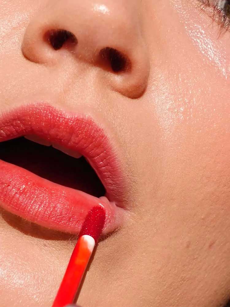 A close-up of a person applying Ere Perez Beetroot Cheek & Lip Tint - Joy 4.5ml with a wand, showing smooth skin and part of the face—ideal for achieving a natural, long-lasting lip stain.