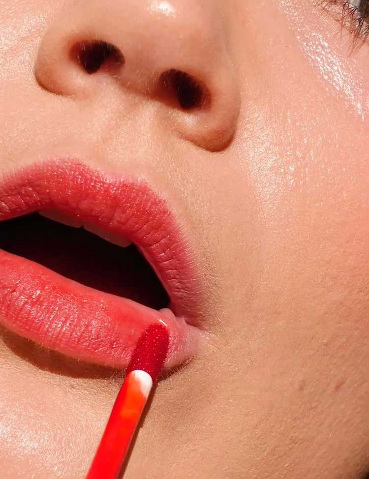 A close-up of a person applying Ere Perez Beetroot Cheek & Lip Tint - Joy 4.5ml with a wand, showing smooth skin and part of the face, ideal for achieving a natural, long-lasting lip stain.