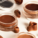 Three open jars of Ere Perez Cacao Bronzing Pot - Playa 7.5g sit among chocolate curls on a light surface, suggesting a sun-kissed glow. A closed silver lid appears in the upper left corner.