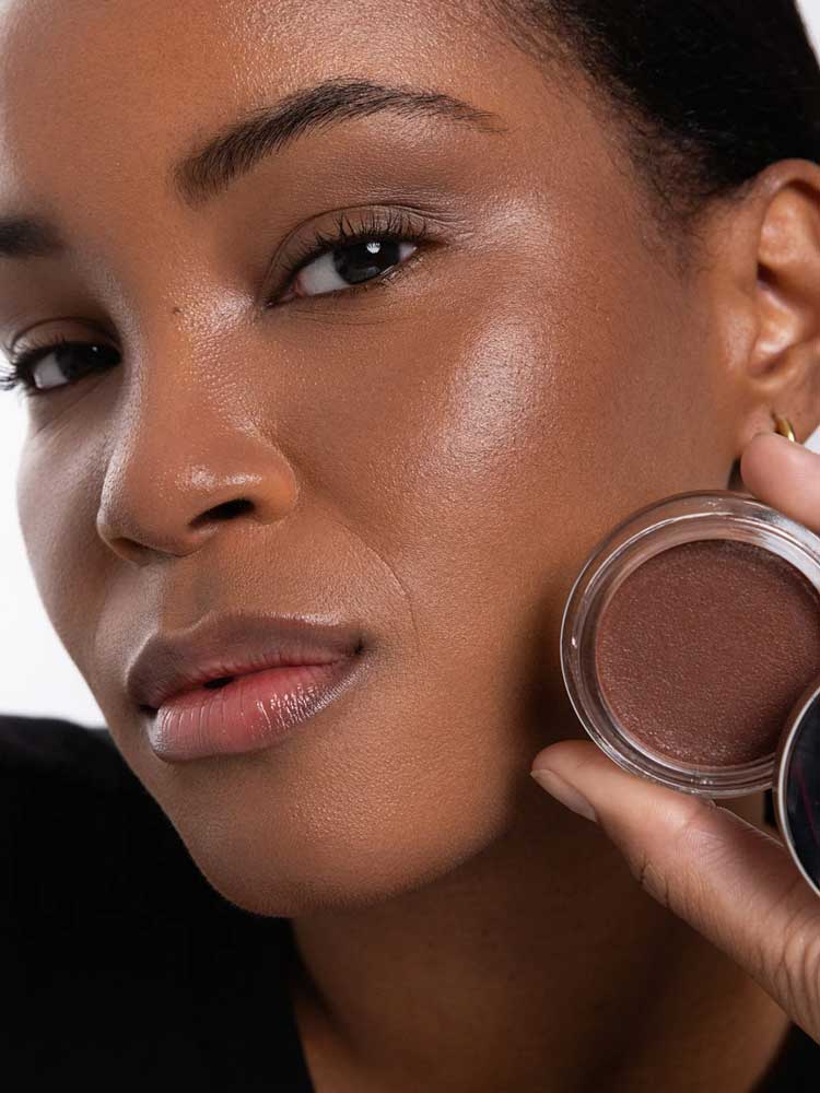 A woman with smooth, glowing brown skin holds an open Ere Perez Cacao Bronzing Pot - Costa 7.5g near her face, highlighting her natural makeup look and radiant complexion.