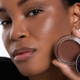 A woman with smooth, glowing brown skin holds an open Ere Perez Cacao Bronzing Pot - Costa 7.5g near her face, highlighting her natural makeup look and radiant complexion.