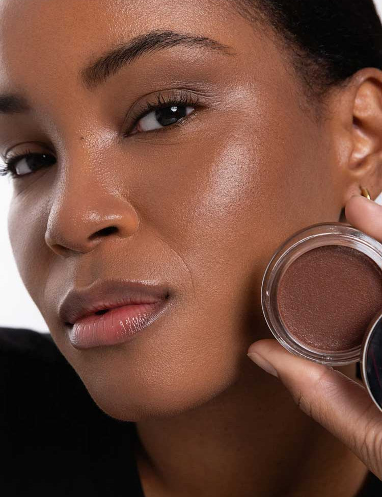 A woman with smooth, glowing brown skin holds an open Ere Perez Cacao Bronzing Pot - Costa 7.5g near her face, highlighting her natural makeup look and radiant complexion.