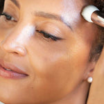 A close-up of a woman applying Ere Perez Cacao Bronzing Pot - Sol 7.5g to her brow bone with a makeup brush, revealing radiant, glowing skin and a natural glow.