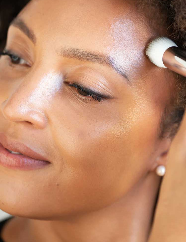 A close-up of a woman applying Ere Perez Cacao Bronzing Pot - Sol 7.5g to her brow bone with a makeup brush, revealing radiant, glowing skin and a natural glow.