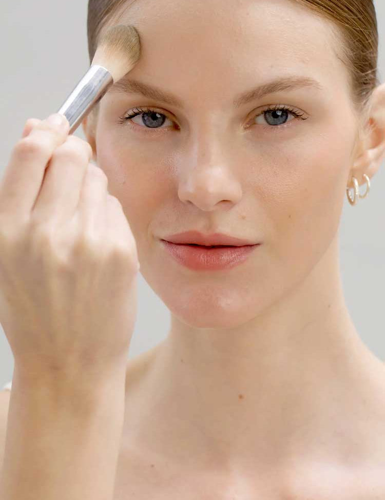 A fair-skinned, light-eyed person with natural makeup and neat brows uses Ere Perez Corn Setting Powder - Original 7g on their forehead with a brush. They look at the camera, have a matte finish, and wear small hoop earrings.