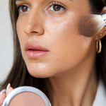 A person applies Ere Perez Rice Powder Blush & Bronzer - Roma 9g to their cheek with a makeup brush while holding an open compact. They have long brown hair, natural makeup, and wear gold hoop earrings.