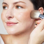A fair-skinned woman with light brown hair applies Ere Perez Rice Powder Blush & Bronzer - Roma Refill 9g to her cheek. She wears a white top and natural makeup, with the vegan refill showcased against a softly lit, neutral background.