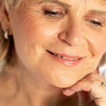 Close-up of an older woman with light skin and short blonde hair, smiling gently with her chin on her hand. She wears small earrings and a ring, her natural glow enhanced by Ere Perez Rice Powder Blush & Bronzer - Brooklyn 9g.