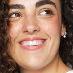 A woman with curly brown hair and clear skin smiles to the side, showing her white teeth. She wears a small pearl earring, a white top, and Ere Perez Rice Powder Blush & Bronzer - Brooklyn 9g for a radiant glow.
