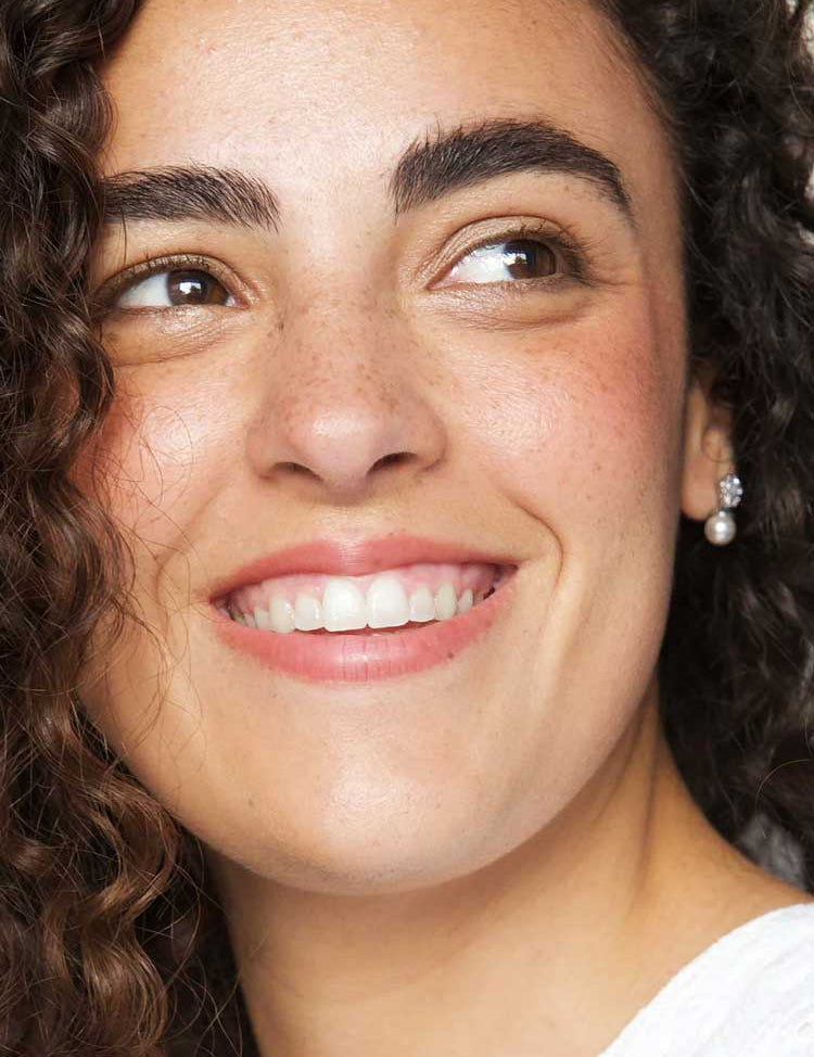 A woman with curly brown hair and clear skin smiles to the side, showing her white teeth. She wears a small pearl earring, a white top, and Ere Perez Rice Powder Blush & Bronzer - Brooklyn 9g for a radiant glow.