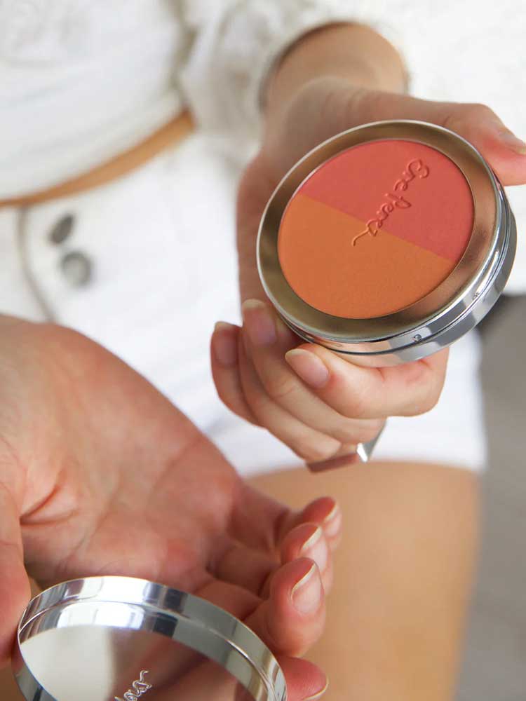 Someone holds an open Ere Perez Rice Powder Blush - Bondi Refill 9g compact featuring three vegan shades—orange, coral, and peach—while dressed in white shorts and a white top.