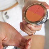 Someone holds an open Ere Perez Rice Powder Blush - Bondi Refill 9g compact featuring three vegan shades—orange, coral, and peach—while dressed in white shorts and a white top.