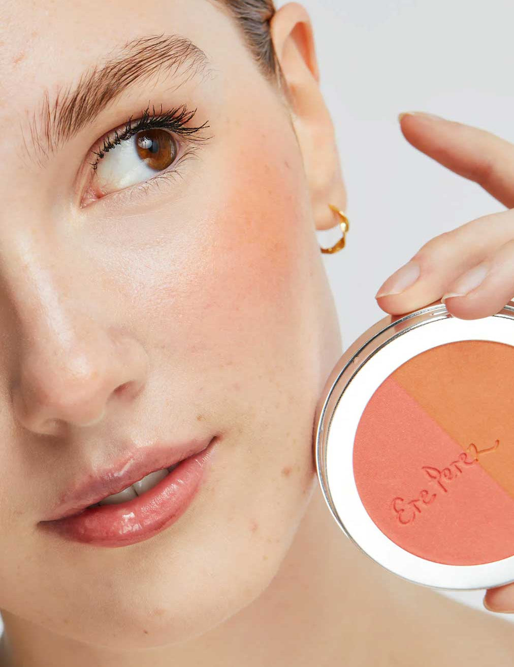 A woman with natural makeup holds the Ere Perez Rice Powder Blush - Bondi Refill 9g near her cheek, wearing a gold hoop and highlighting the vegan peach, pink, and tan shades inspired by Bondi beauty.