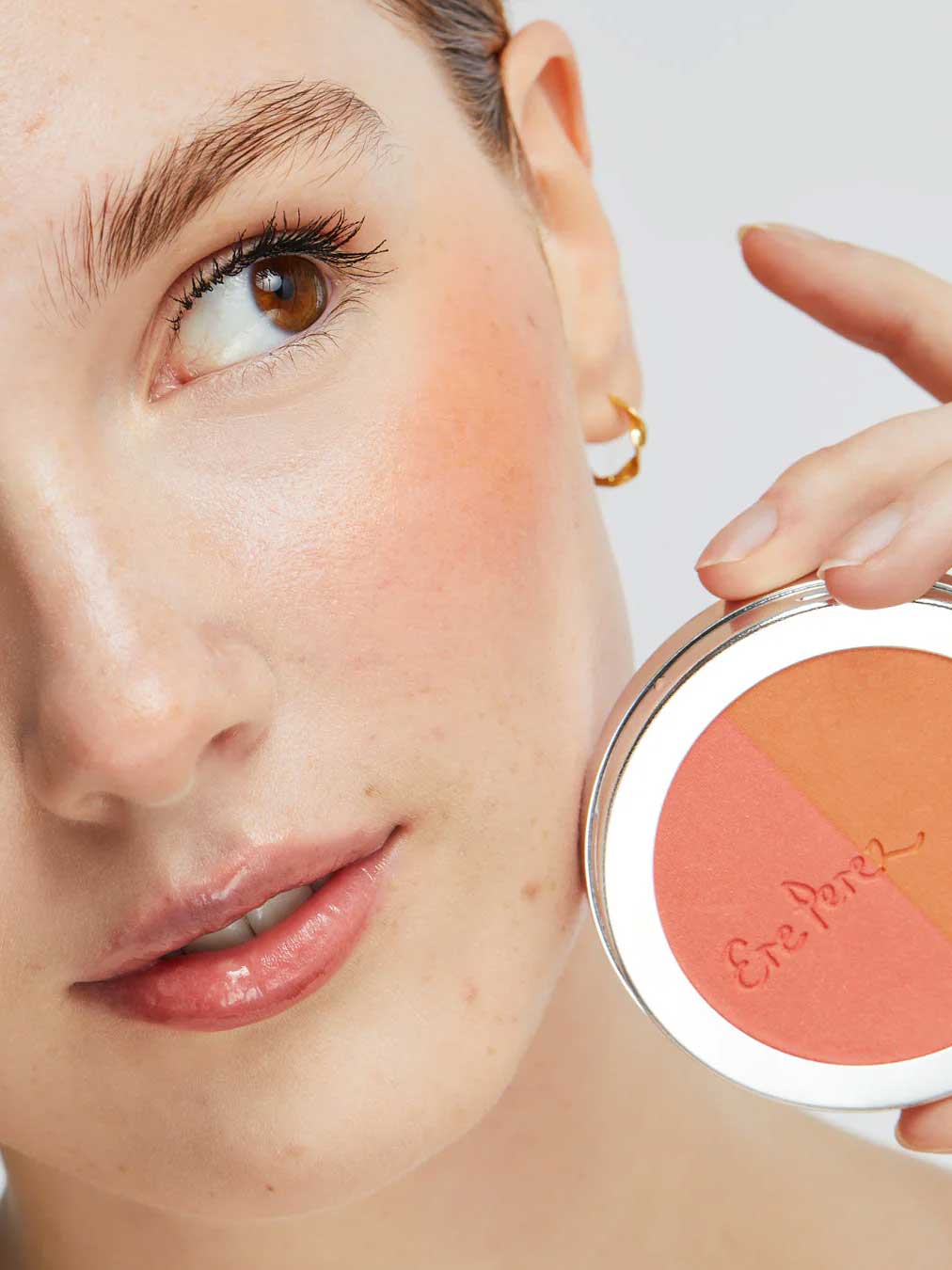 A woman with natural makeup holds the Ere Perez Rice Powder Blush - Bondi Refill 9g near her cheek, wearing a gold hoop and highlighting the vegan peach, pink, and tan shades inspired by Bondi beauty.