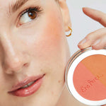 A woman with natural makeup holds the Ere Perez Rice Powder Blush - Bondi Refill 9g near her cheek, wearing a gold hoop and highlighting the vegan peach, pink, and tan shades inspired by Bondi beauty.