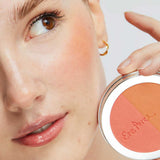 A woman with natural makeup holds the Ere Perez Rice Powder Blush - Bondi Refill 9g near her cheek, wearing a gold hoop and highlighting the vegan peach, pink, and tan shades inspired by Bondi beauty.