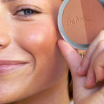 A woman smiles while holding a compact with two blush shades and Ere Perez Rice Powder Bronzer - Tulum 9g near her cheek, displaying a soft, glowing look achieved with this natural bronzer.