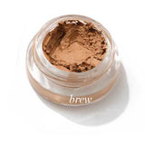 A small clear jar of Ere Perez Arnica All-Cover Pot 5g in creamy, light brown “brew” rests on a white background, its smooth, buildable concealer surface slightly scooped out.