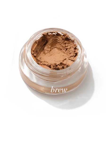 A small clear jar of Ere Perez Arnica All-Cover Pot 5g in creamy, light brown “brew” rests on a white background, its smooth, buildable concealer surface slightly scooped out.