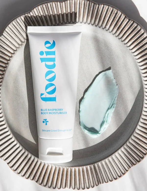 Foodie Blue Raspberry Body Moisturiser tube on a ridged silver plate beside a thick pale aqua cream smear, with striped shadows across the scene.