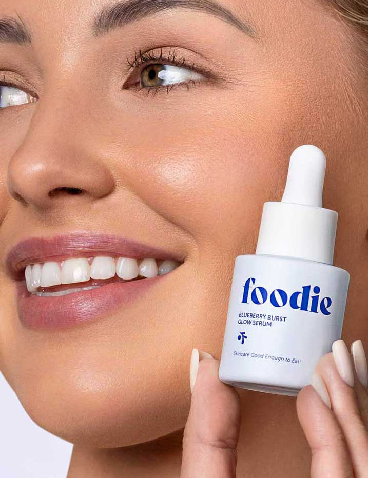 A smiling person holds Foodie Blueberry Burst Glow Serum 20ml near their face, showing off smooth, glowing skin. Ideal skincare for teens, the image highlights their radiant complexion and the featured serum.