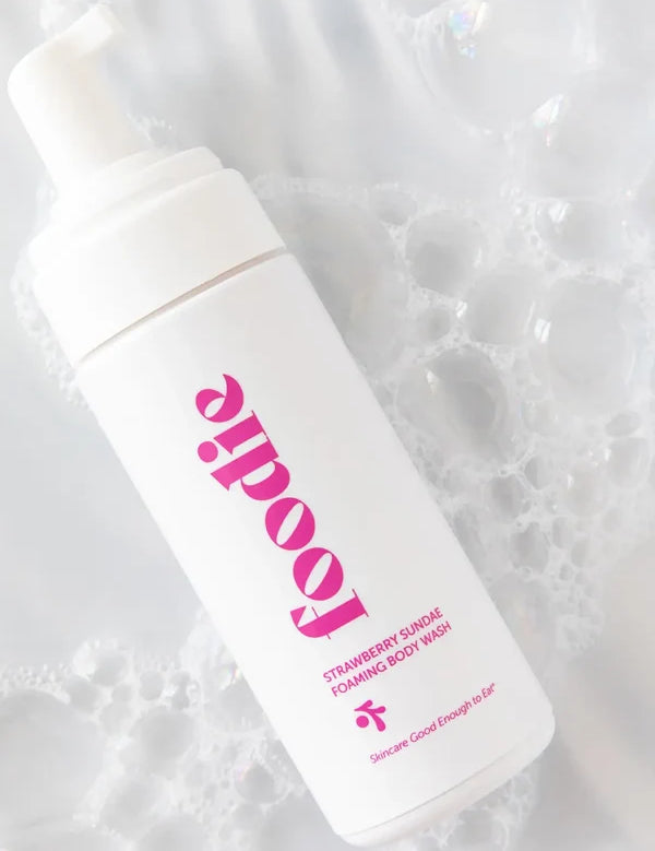 White Foodie Strawberry Sundae Foaming Body Wash bottle with pink lettering lying diagonally among white soap bubbles, with the pump exposed.