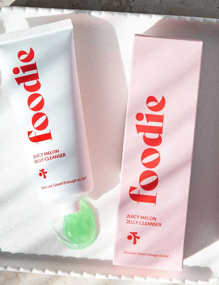 A white tube of Foodie Juicy Melon Jelly Cleanser 75ml dispenses green gel beside its pink box on a white textured surface, with sunlight creating soft shadows.