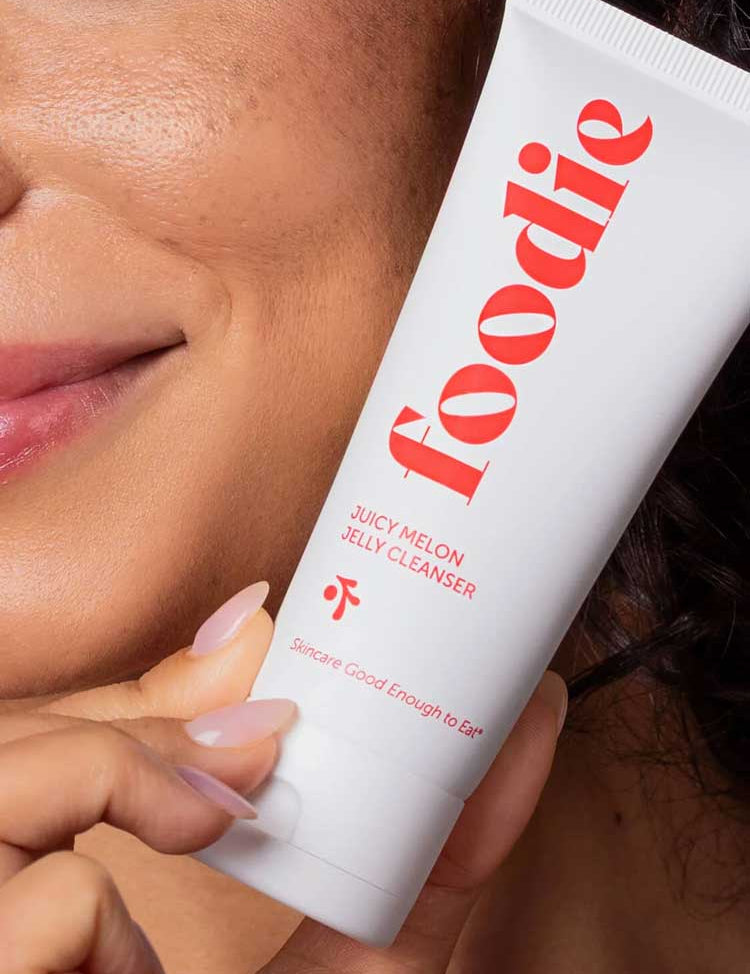 A person with smooth skin smiles, holding a white tube of Foodie Juicy Melon Jelly Cleanser 75ml—a gentle natural face wash—close to their face. The tube reads: Skincare Good Enough to Eat.