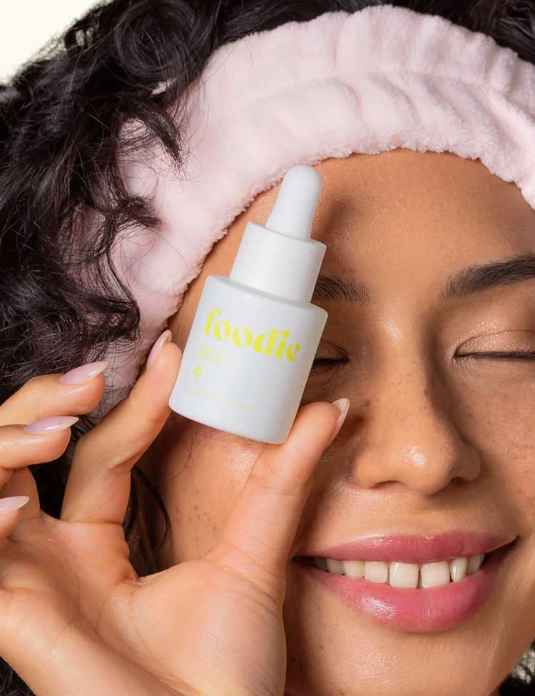 A smiling woman with curly hair and a pink headband holds Foodie Lemon Aid Pimple Gel 20ml close to her face, eyes closed, enjoying this natural spot treatment for teenage skin.