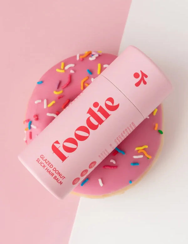 Closed pink Foodie Glazed Donut Slick Hair Balm tube lying diagonally across a pink-iced doughnut with colourful sprinkles, on a pink-and-white background.