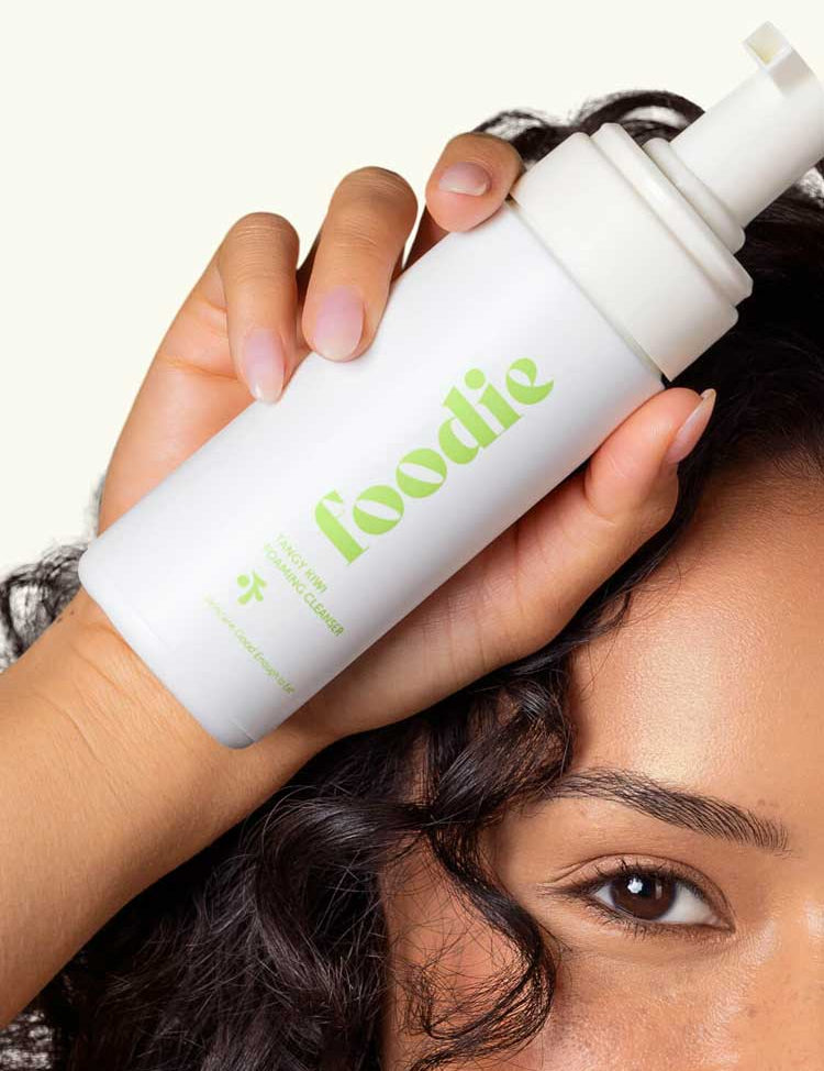 A person holds a white bottle labeled "Foodie Tangy Kiwi Foaming Facial Cleanser 150ml" near their face; the close-up shows one eye, eyebrow, and curly hair, highlighting this natural face wash for teens.
