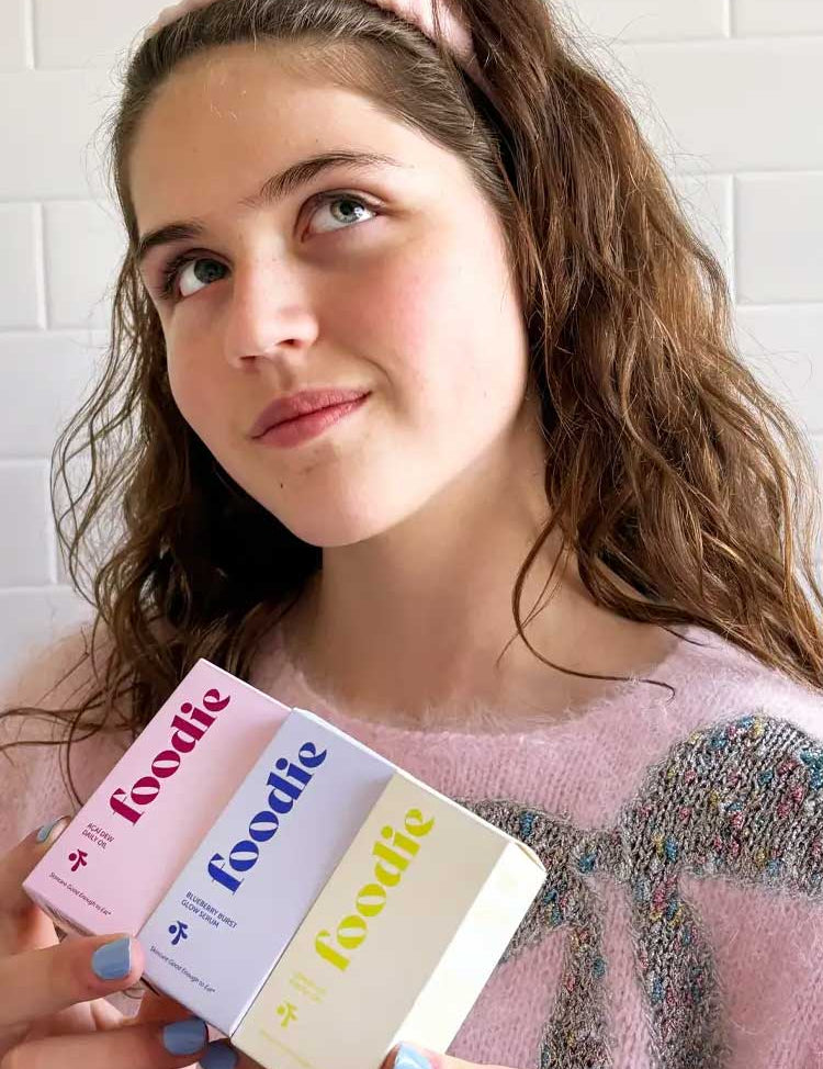 A young woman with wavy brown hair and a pink headband holds three colorful boxes labeled Foodie Açai Dew Daily Oil 20ml. Dressed in a light pink sweater, she stands before white tiles, looking upward thoughtfully.