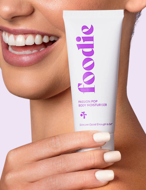 A smiling woman holds a white Foodie Passion Pop Body Moisturiser tube beside her cheek, with its large purple lettering facing the camera.