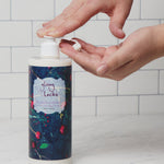 One hand presses the pump of a 100% Pure Glossy Locks Repair Conditioner bottle, dispensing white conditioner into the other open palm. The bottle has a blue fruit-patterned label; white tiles are behind it.