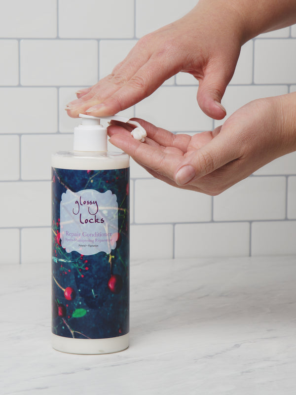 One hand presses the pump of a 100% Pure Glossy Locks Repair Conditioner bottle, dispensing white conditioner into the other open palm. The bottle has a blue fruit-patterned label; white tiles are behind it.